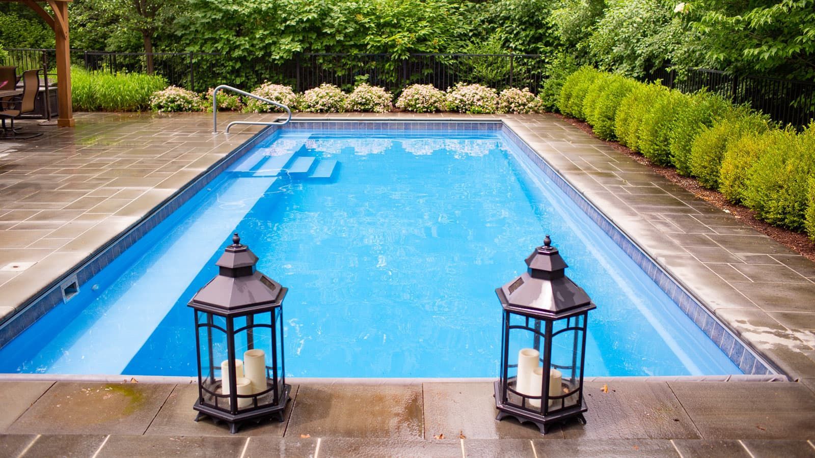 Blue swimming pool with two black lanterns on a stone patio, surrounded by greenery