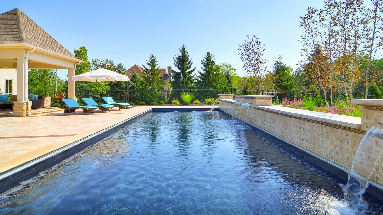 Backyard infinity pool beside patio lounge chairs and a pavilion, with trees and blue sky beyond