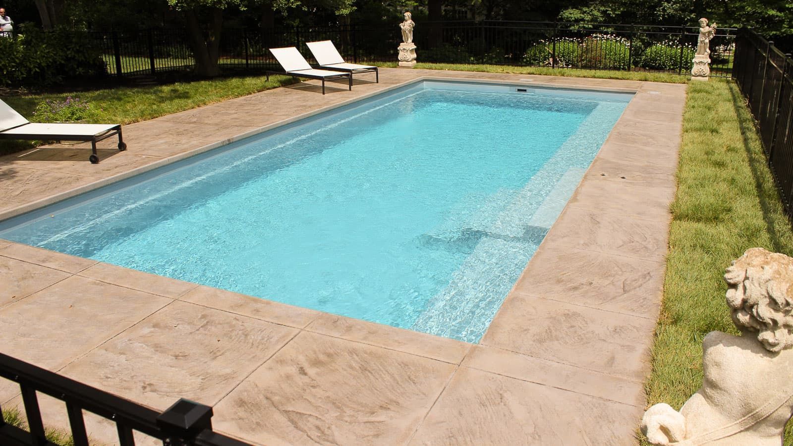 Rectangular backyard swimming pool with blue water, tan deck, and lounge chairs in a garden setting