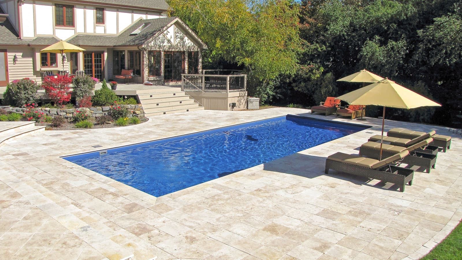 Backyard patio with a blue pool, lounge chairs, umbrellas, and a house in the background