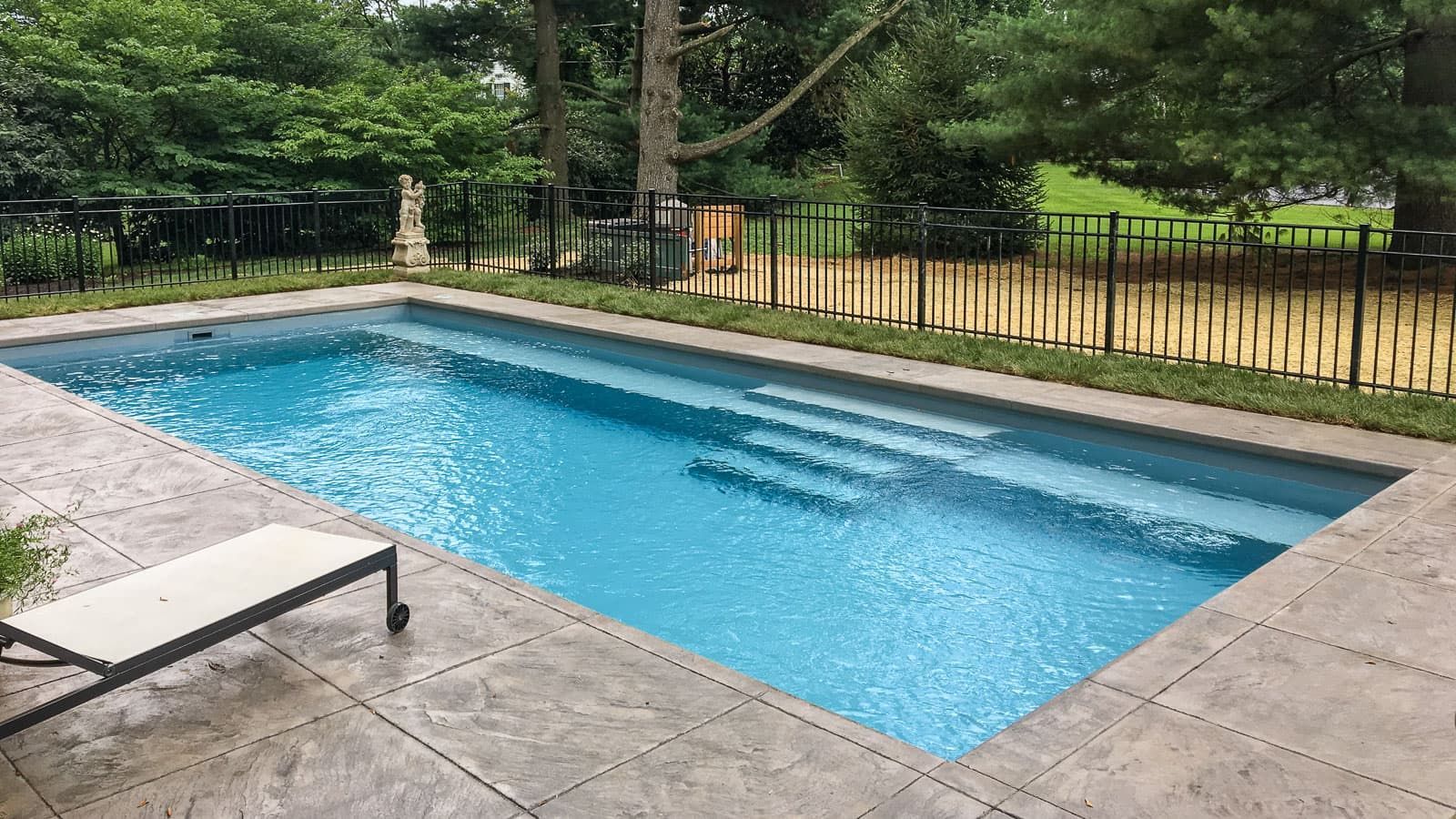 Rectangular outdoor swimming pool with blue water, steps, stone deck, and lounge chair beside a fenced yard