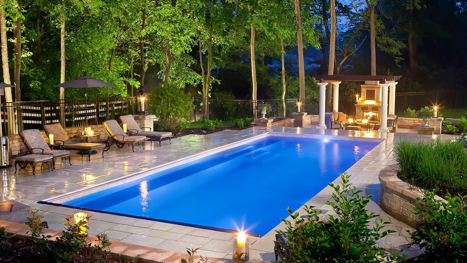 Illuminated backyard pool at night with lounge chairs, palm trees, and a small gazebo