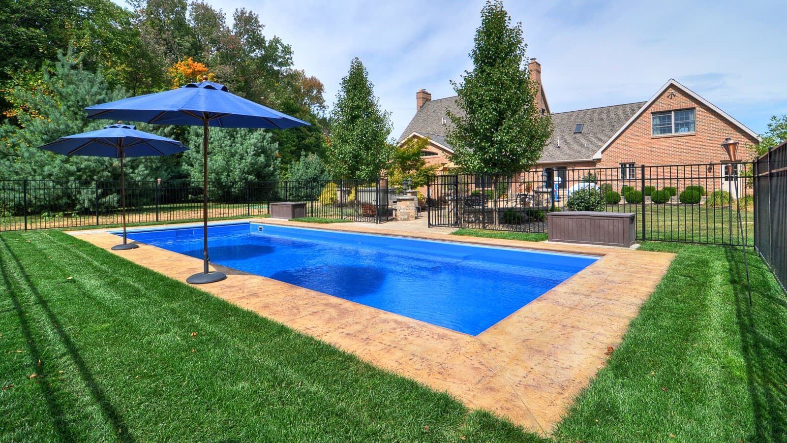 Backyard pool with blue umbrellas, green lawn, and a brick house on a sunny day