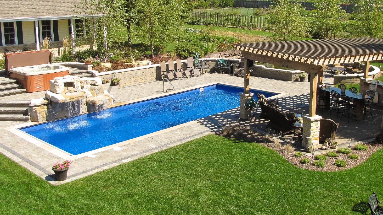 Backyard with a blue swimming pool, stone patio, and covered seating area surrounded by green lawn.