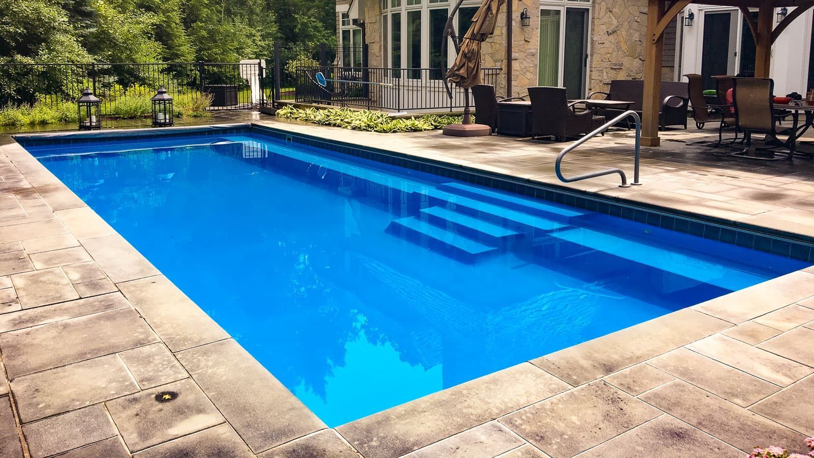 Blue rectangular outdoor swimming pool with stone deck and lounge chairs in the background