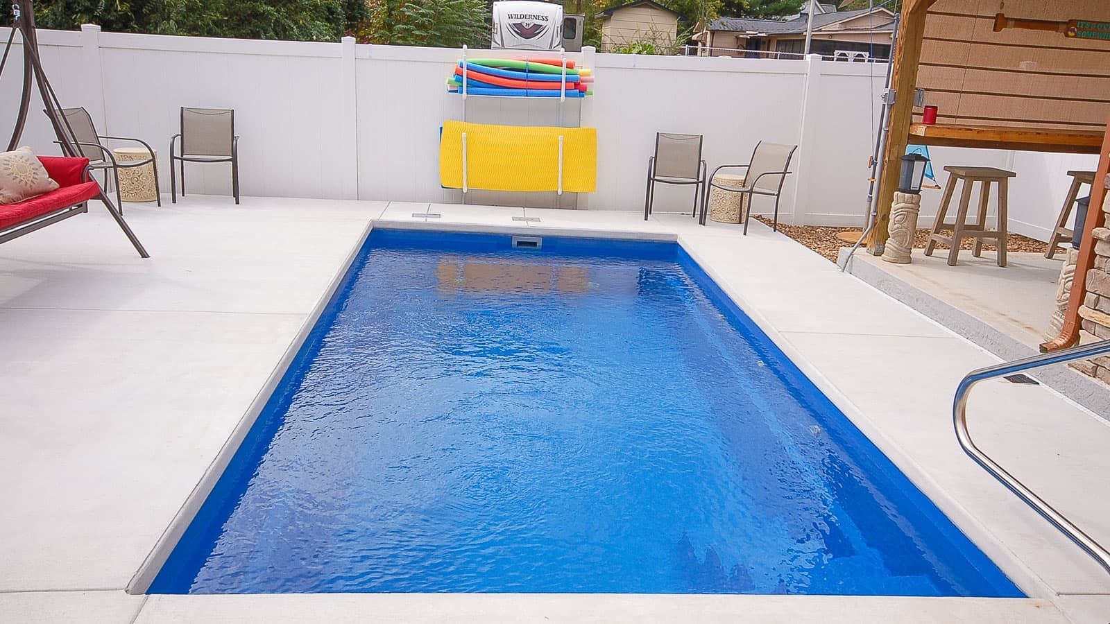 Rectangular blue swimming pool in a white patio with lounge chairs and a small table by the fence
