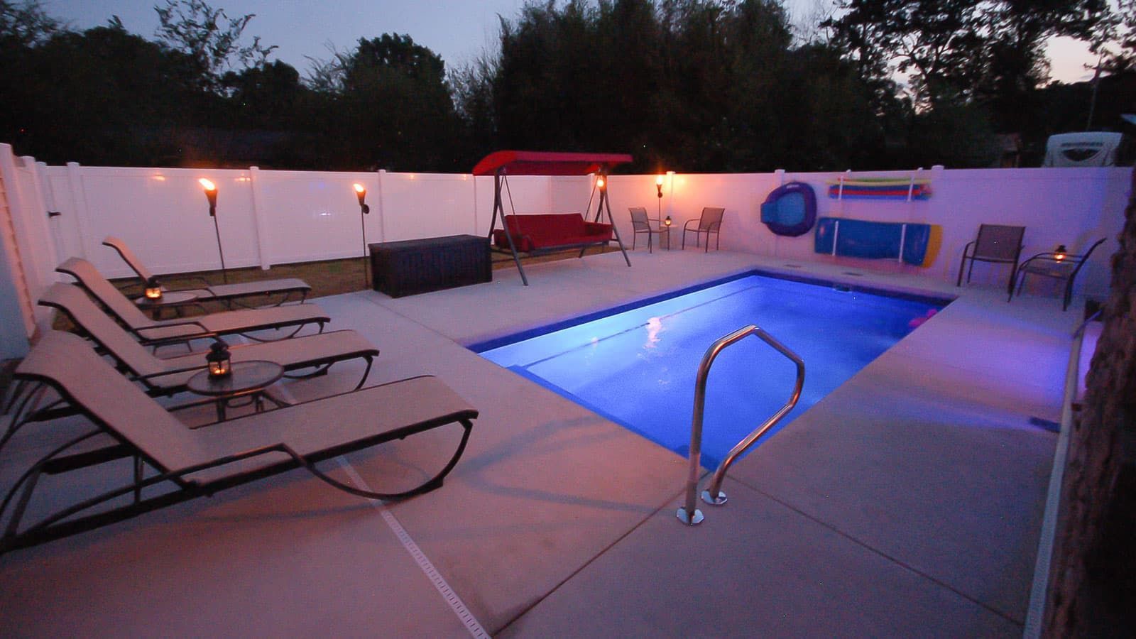 Lit backyard pool with lounge chairs, red canopy, and blue pool lights at dusk