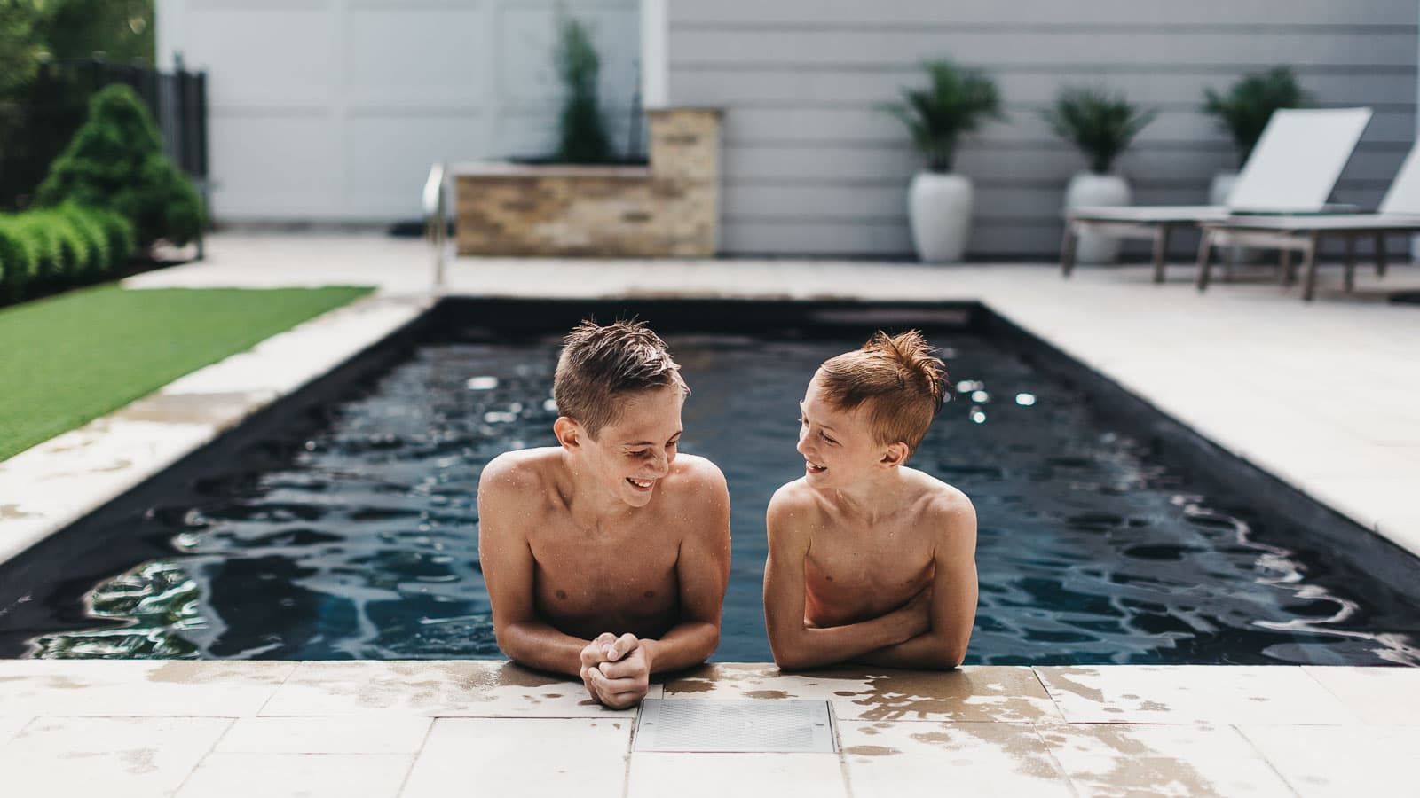 Two children resting at the edge of a backyard pool, with a modern patio and lounge chairs behind them