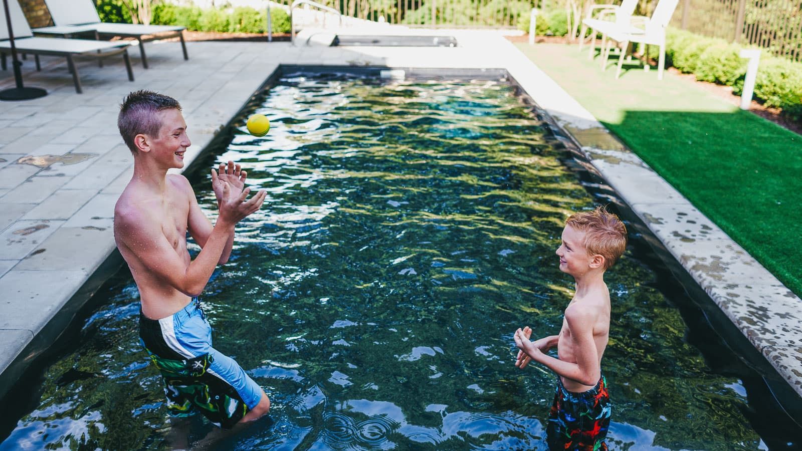 Two boys playing in a backyard pool, splashing water near a floating ball