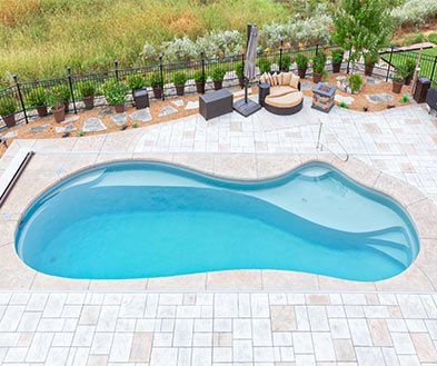 Curved backyard swimming pool with light stone patio, lounge chairs, and garden greenery surrounding it