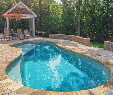 Curved backyard swimming pool with stone deck, lounge chairs, and red umbrella near a wooded fence.