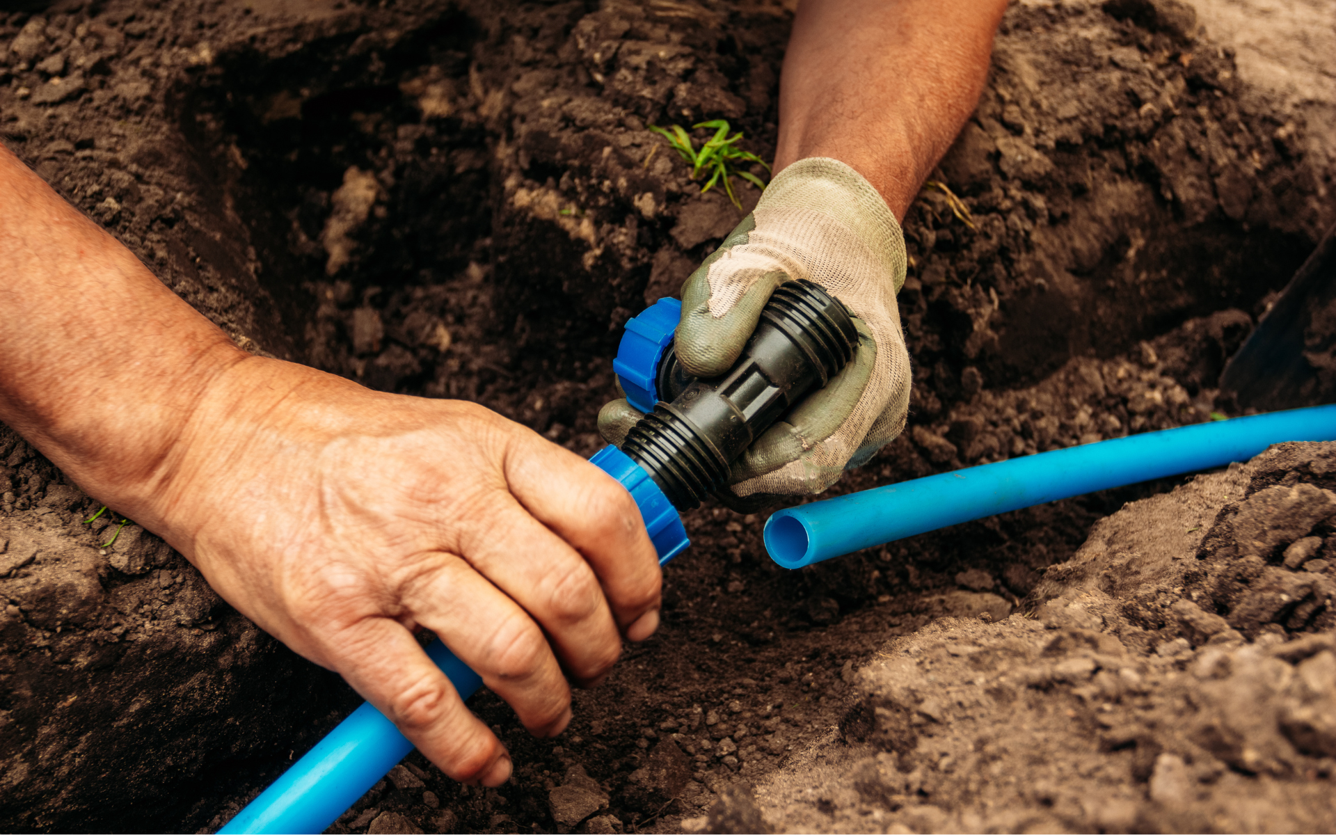 Hands connect a blue pipe fitting in a dirt trench, with one gloved and one bare hand.