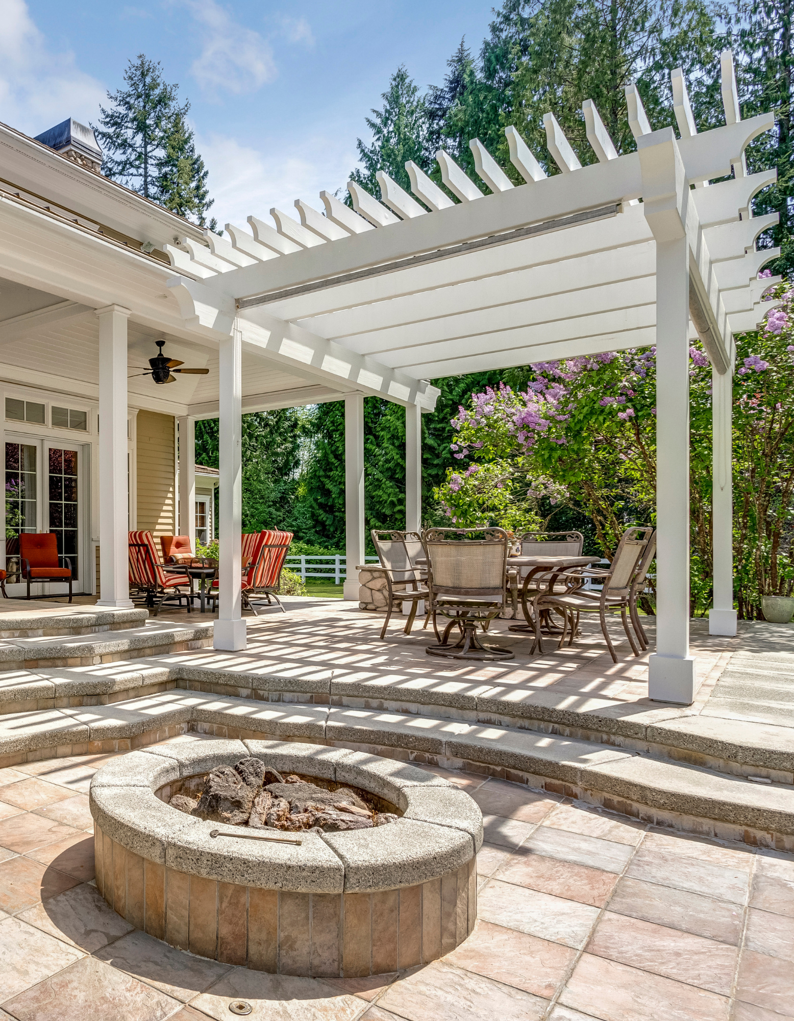 Backyard patio with white pergola, stone fire pit, and outdoor seating in sunlight