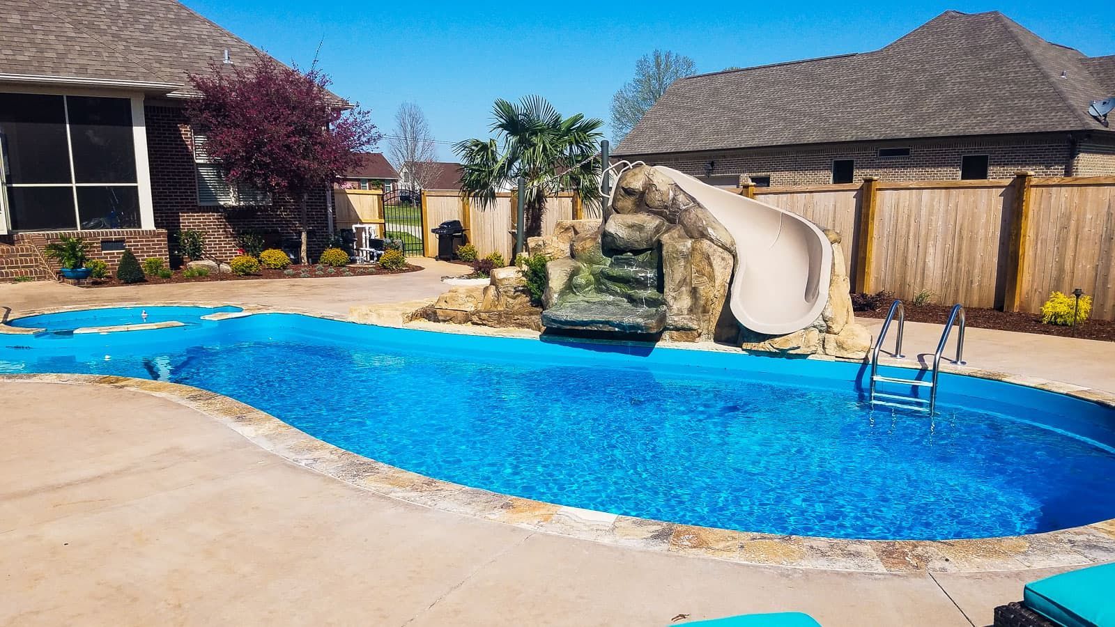 Backyard pool with blue water, slide, and patio chairs beside a house on a sunny day