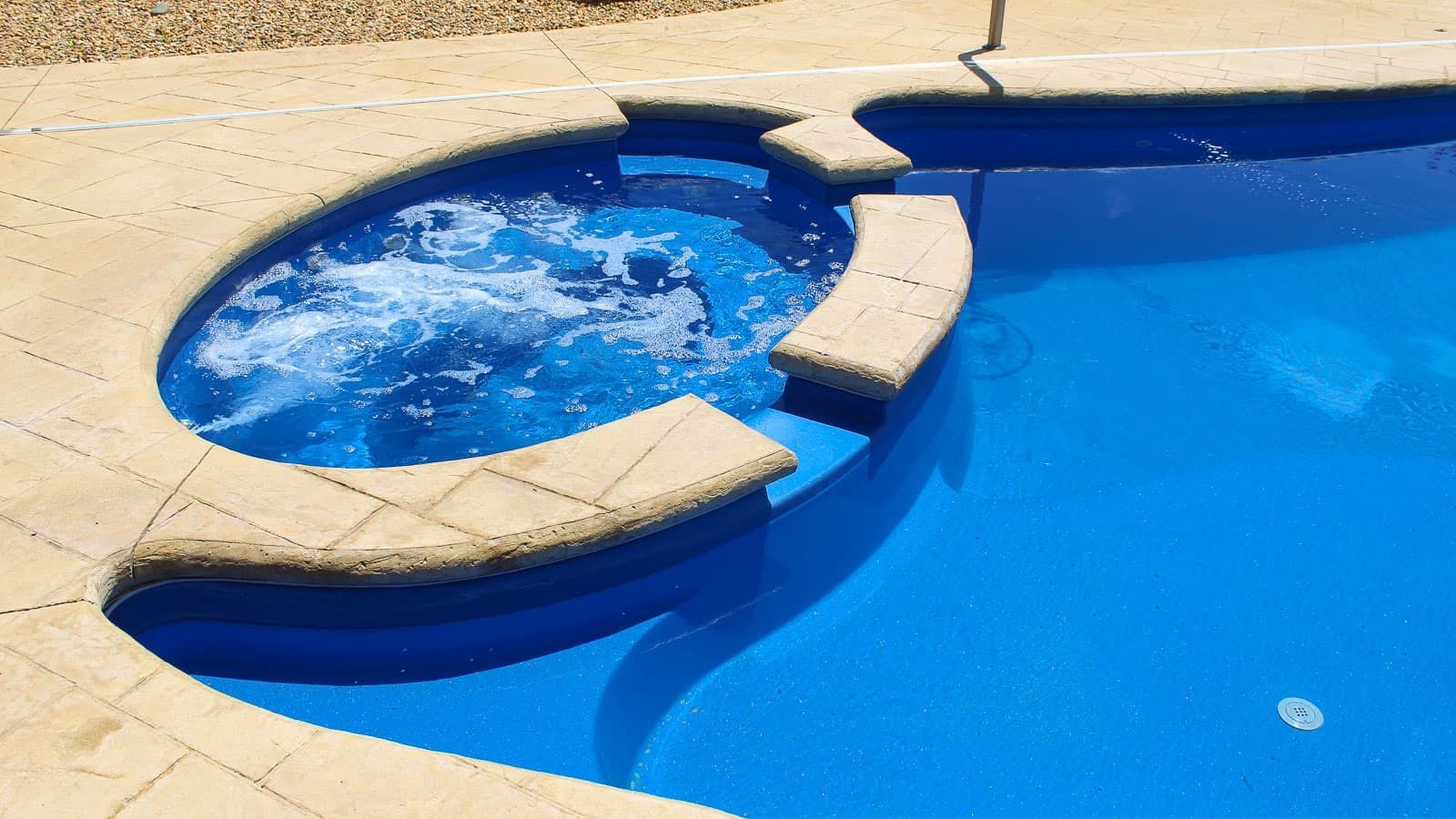 Blue swimming pool with a small round hot tub and beige stone deck tiles