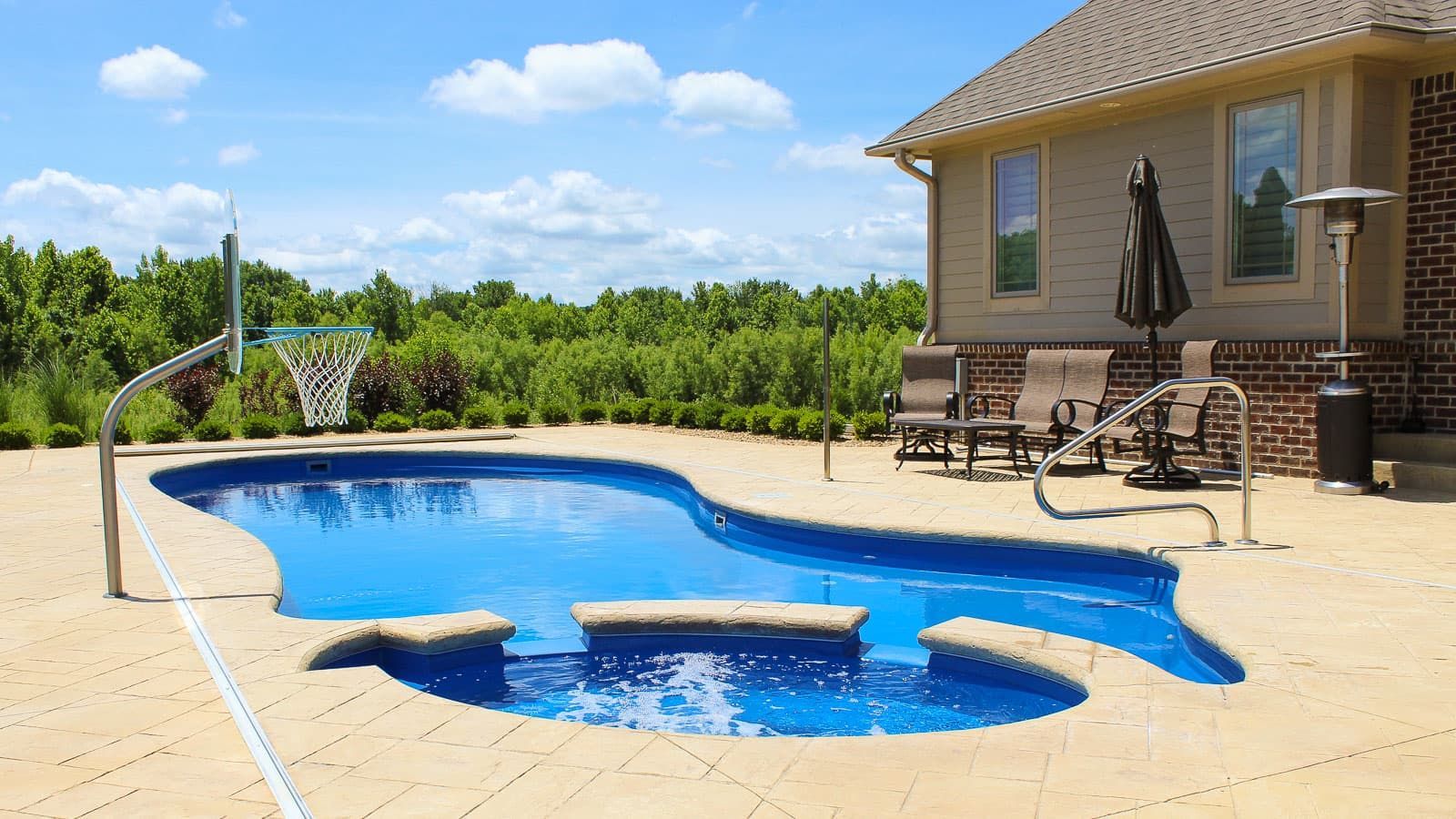 Backyard swimming pool with a tan patio, hot tub, lounge chairs, and a house beside green trees.