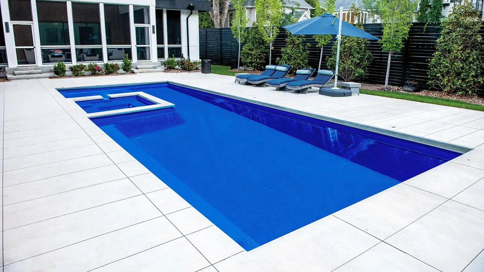 Modern backyard swimming pool with white tile deck, lounge chairs, and blue umbrellas