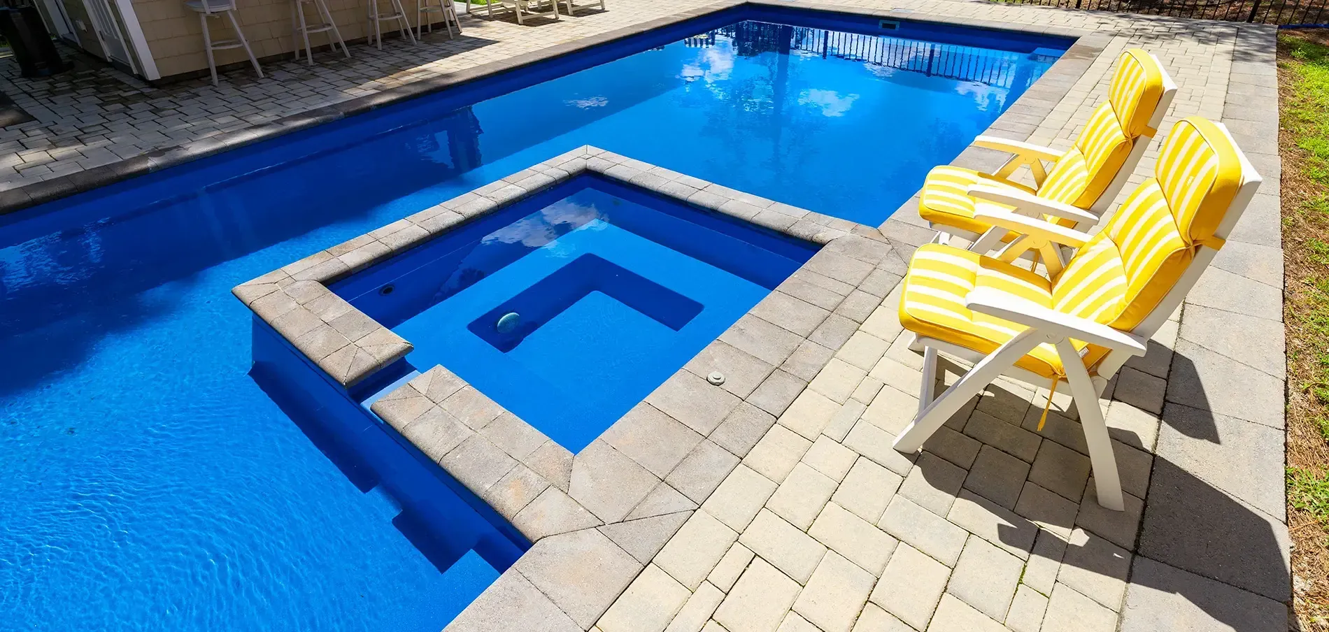 Backyard pool with a small square spa, blue water, and yellow lounge chairs on a tiled deck