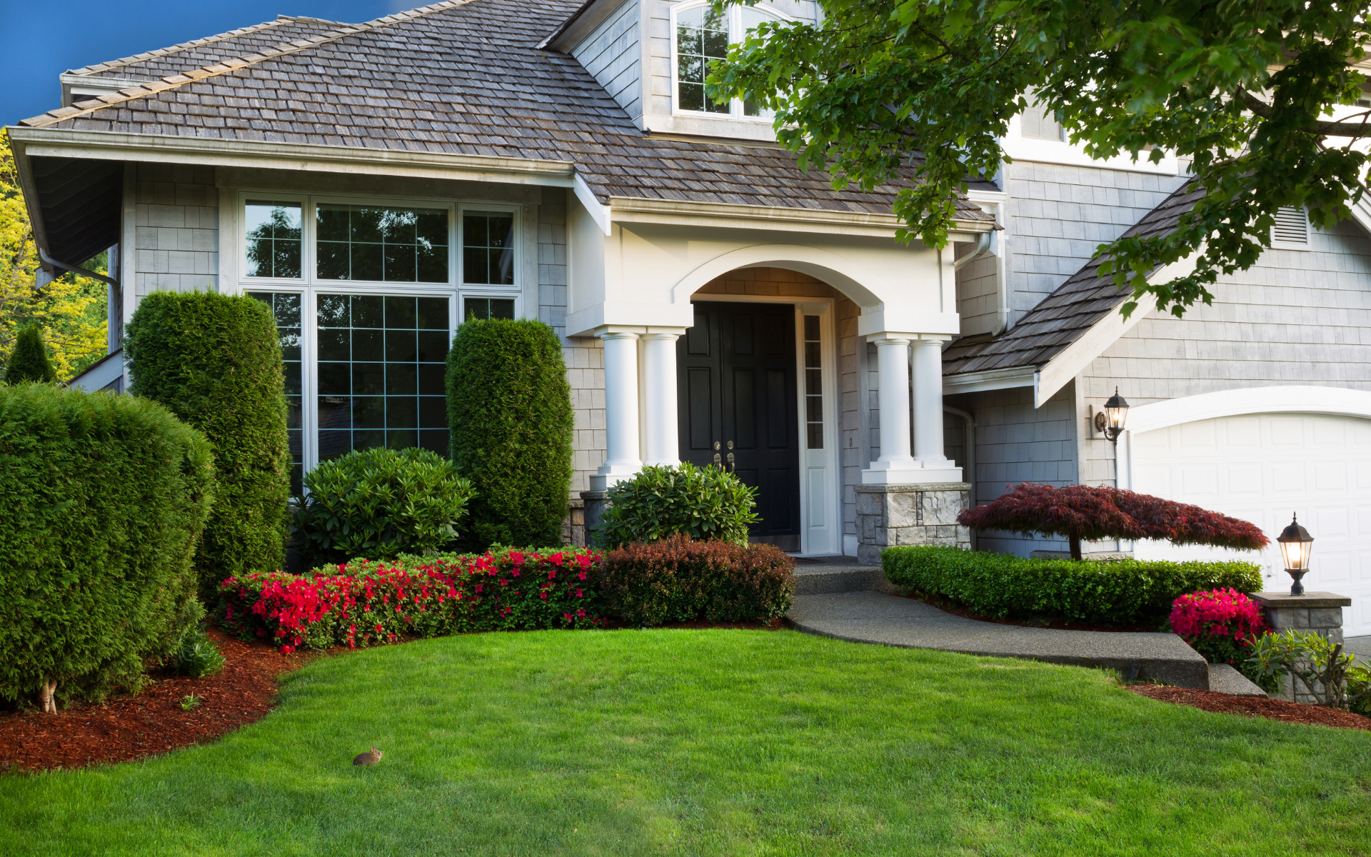 Suburban house with white columns, manicured lawn, trimmed hedges, and red flower beds