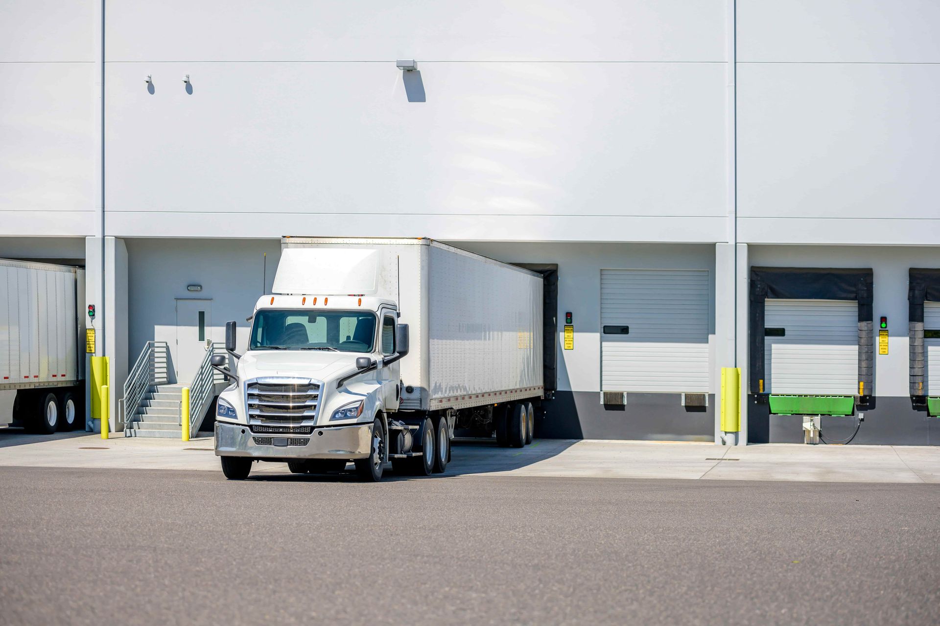 Semi-truck at a loading dock; white truck with trailer backed up to a white warehouse building.