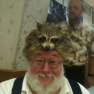 A smiling person wearing a raccoon skin hat, with another person blurred in the background.