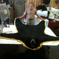A person stands behind a large black bear rug with a gold trim spread out on a white table in a workshop.