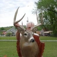 A person holds a large mounted deer trophy with wide antlers against a grassy outdoor residential background.
