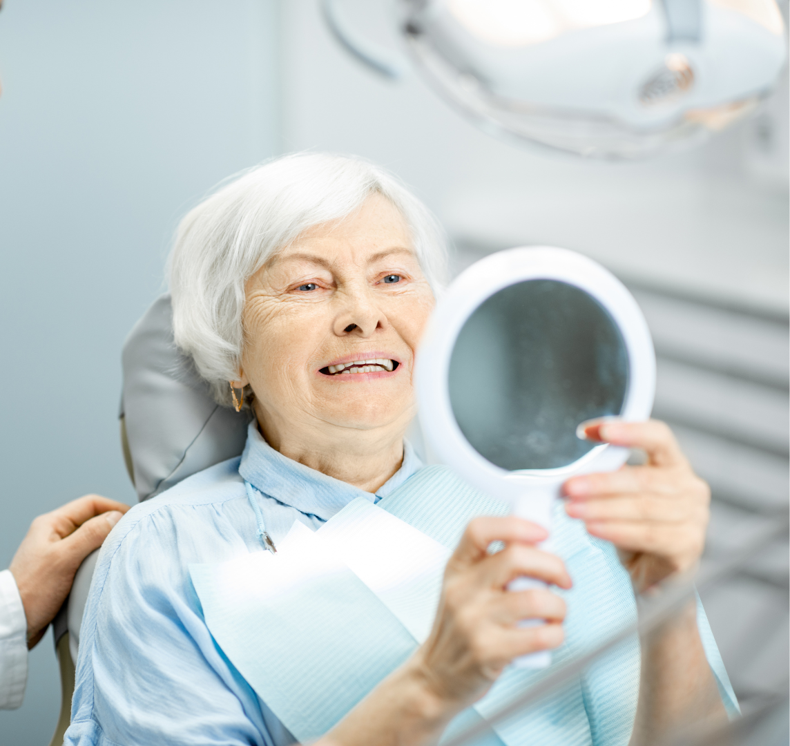 An elderly woman is looking at her teeth in a mirror.