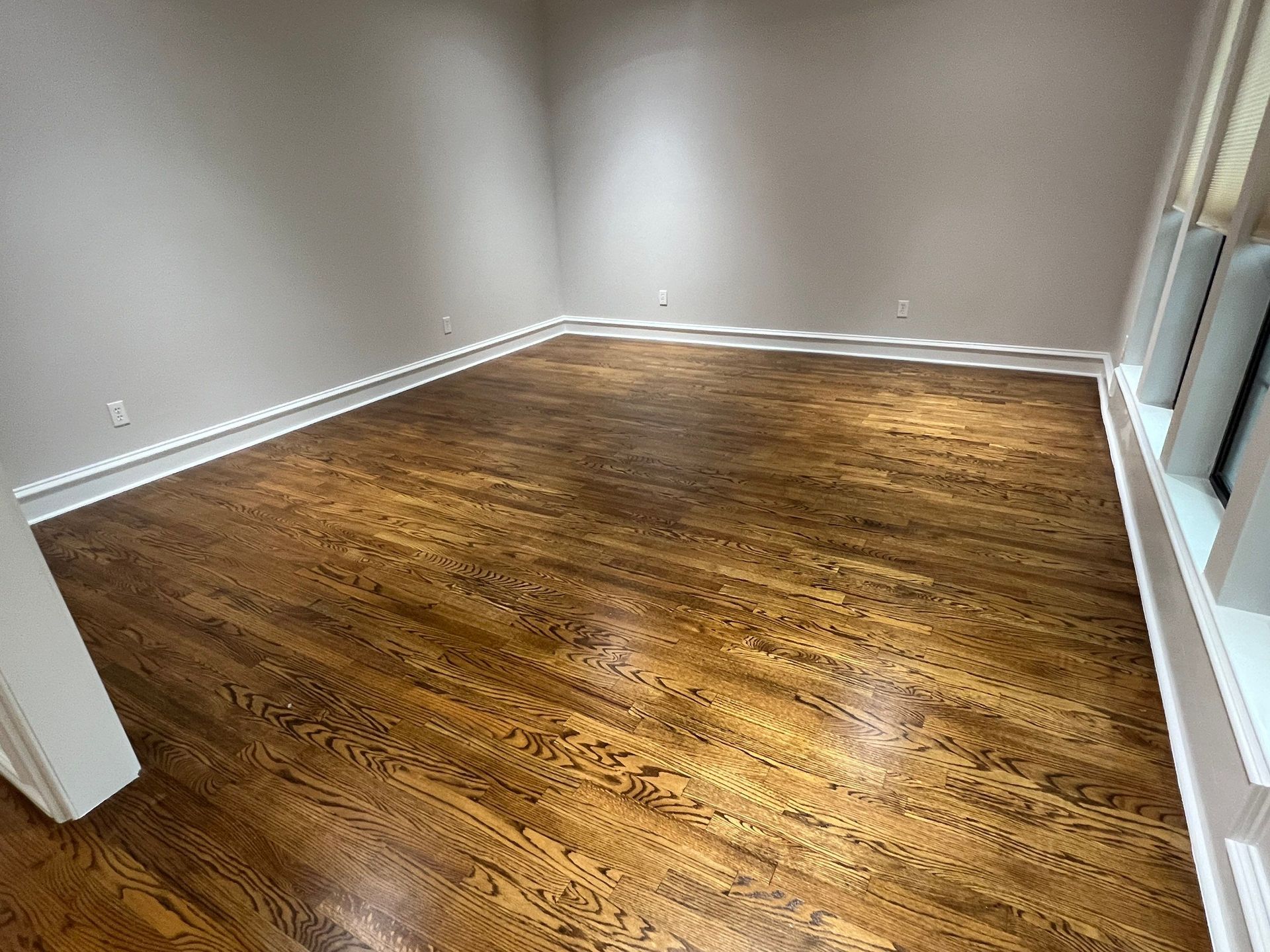 Empty room with stained wood floors and beige walls. White trim outlines the room.