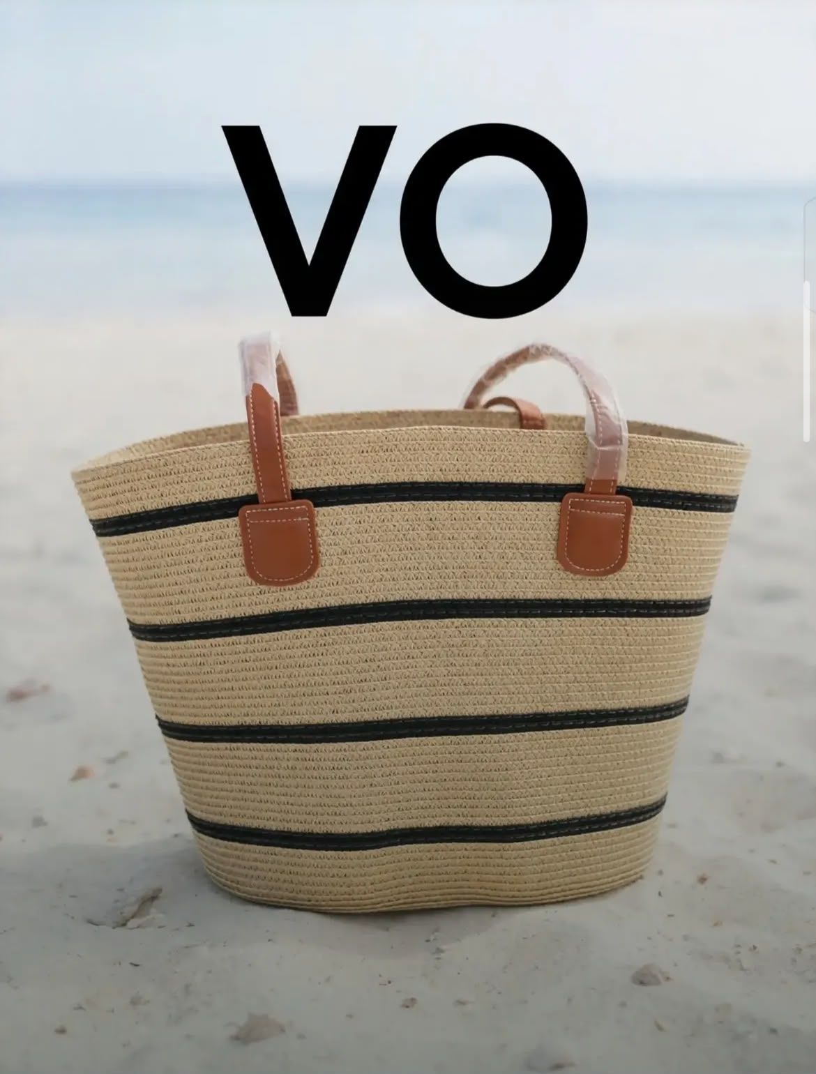 Bolso tote de paja para la playa, con rayas negras y asas color tostado, sobre una superficie arenosa, con «VO» encima
