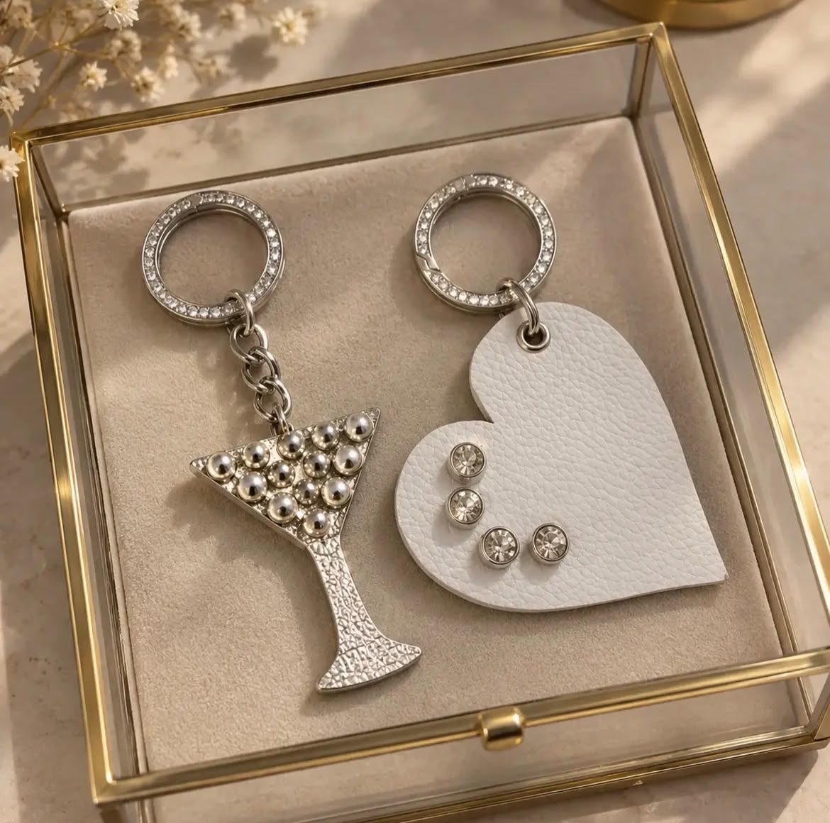 Dos dijes para llavero en una caja con bordes dorados: una copa de martini con pedrería y un corazón blanco con gemas.