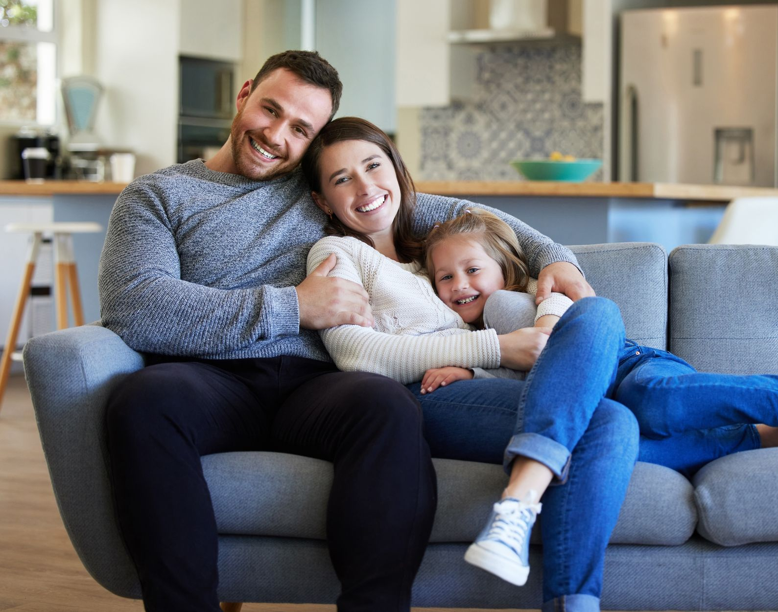 Family relaxing together on a gray couch in a bright living room, smiling and cuddling