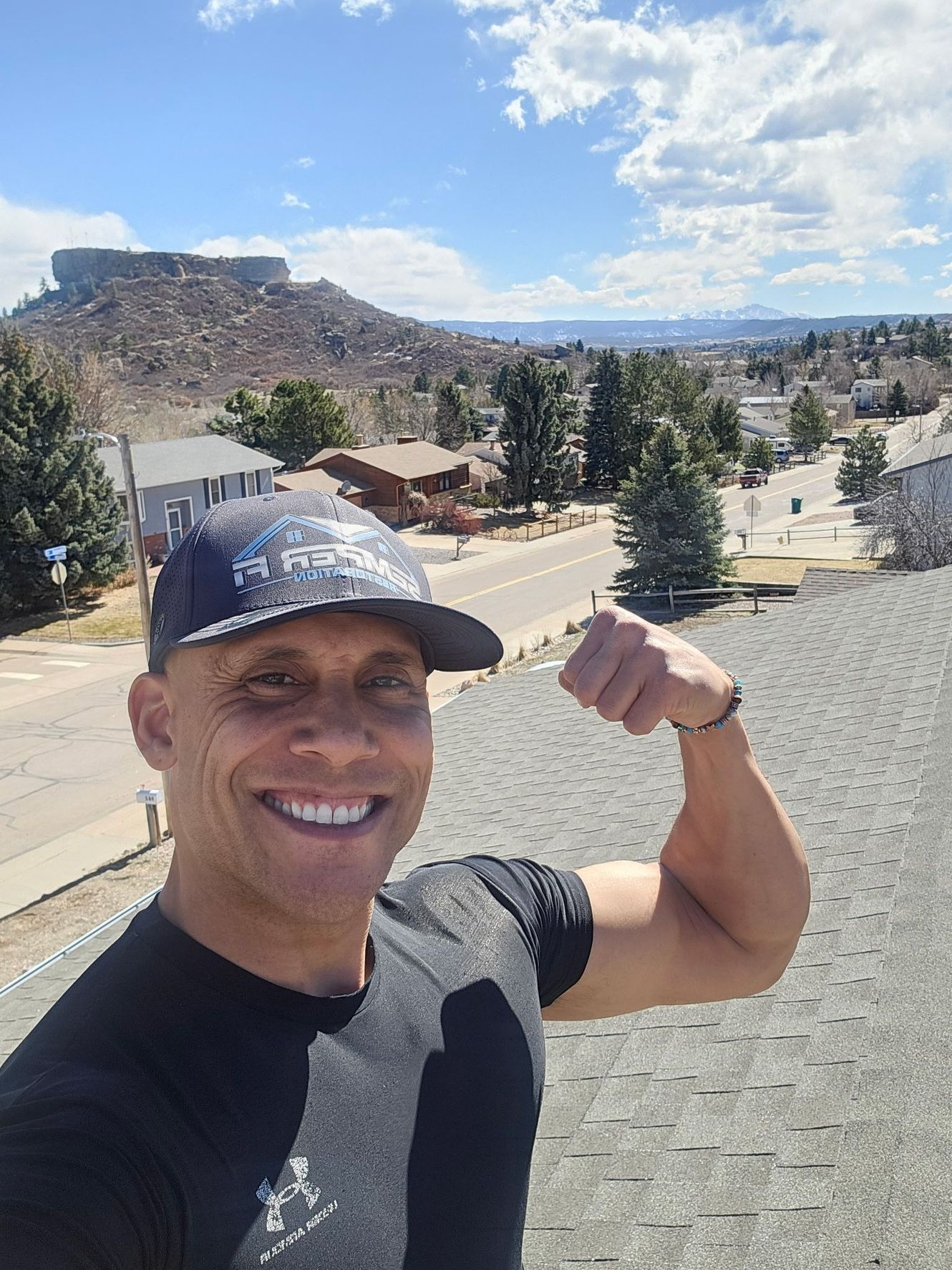 A man wearing a hat is flexing his arm in front of a mountain.