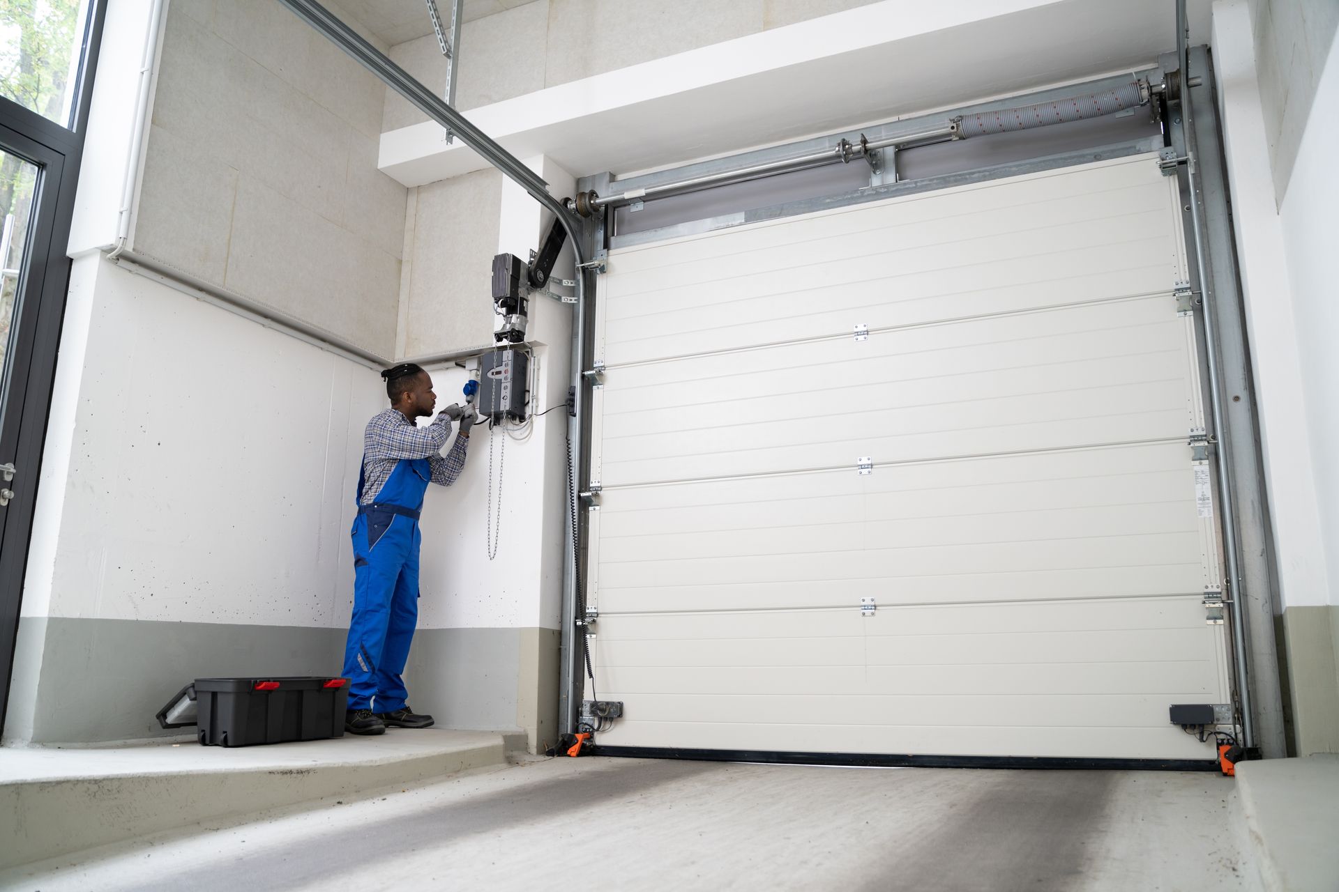 A technician in blue work overalls repairs an electronic control panel next to a large white sectional garage door.