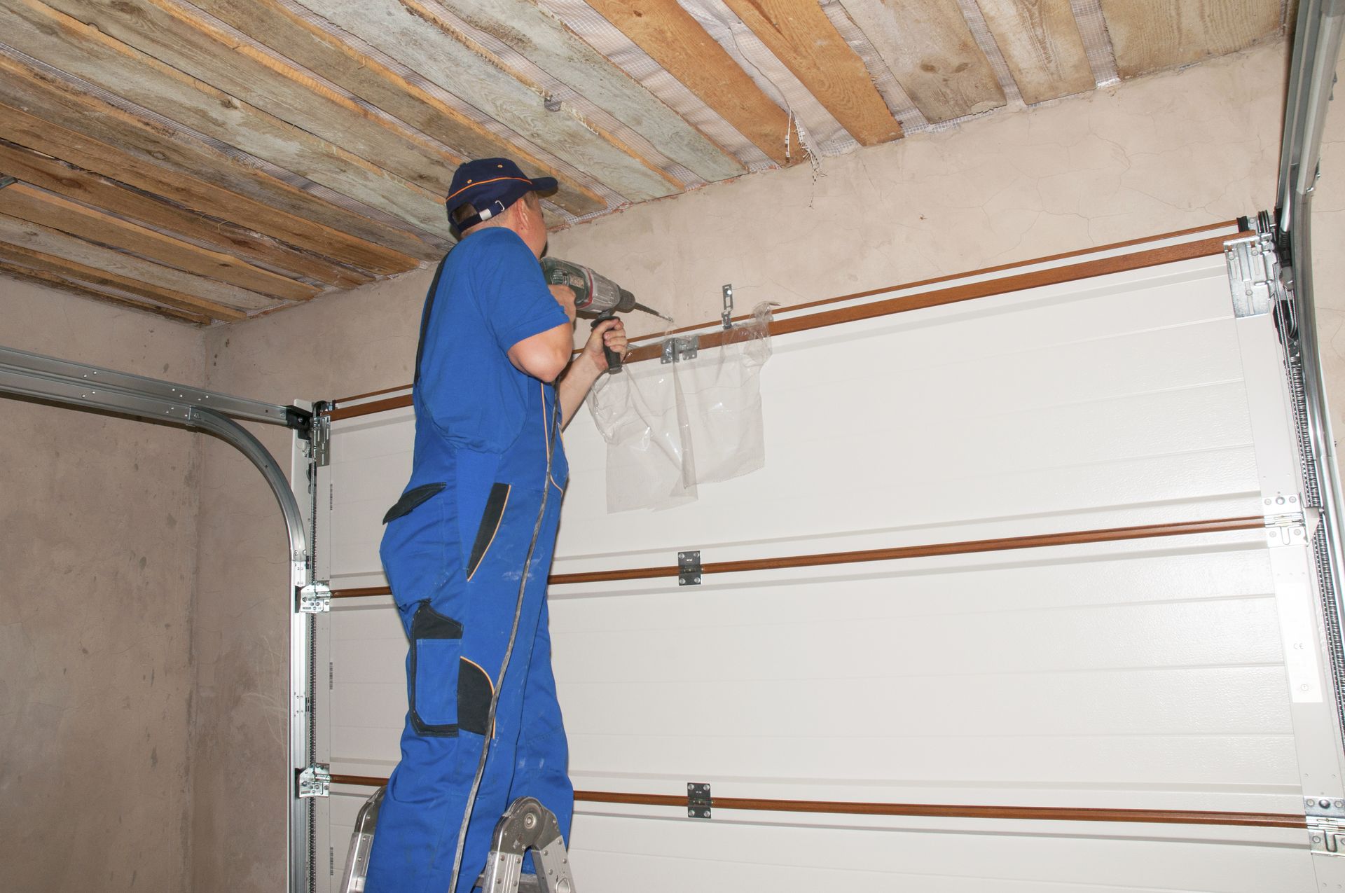 A worker in a blue jumpsuit stands on a ladder, using a power drill to install a white sectional garage door.
