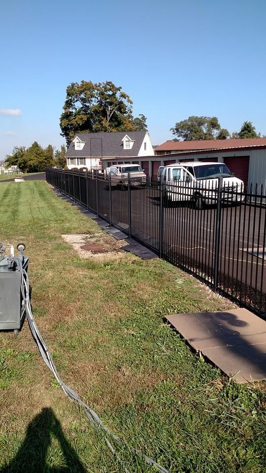 Black metal fence along a grassy area, with a house, cars, and van visible in the background on a sunny day.