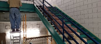 Person on a ladder painting near a green and blue staircase with brick-like objects. Tile walls.