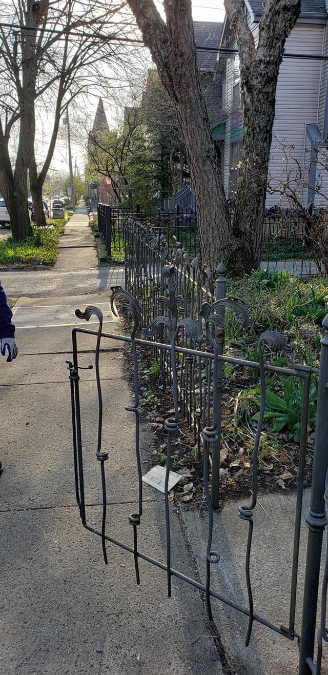 Metal fence in front of a house, sidewalk in foreground. Trees and a person are in the background.