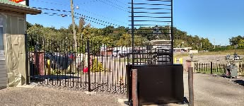A black metal gate, trees, and a building on a gravel lot under a bright blue sky.