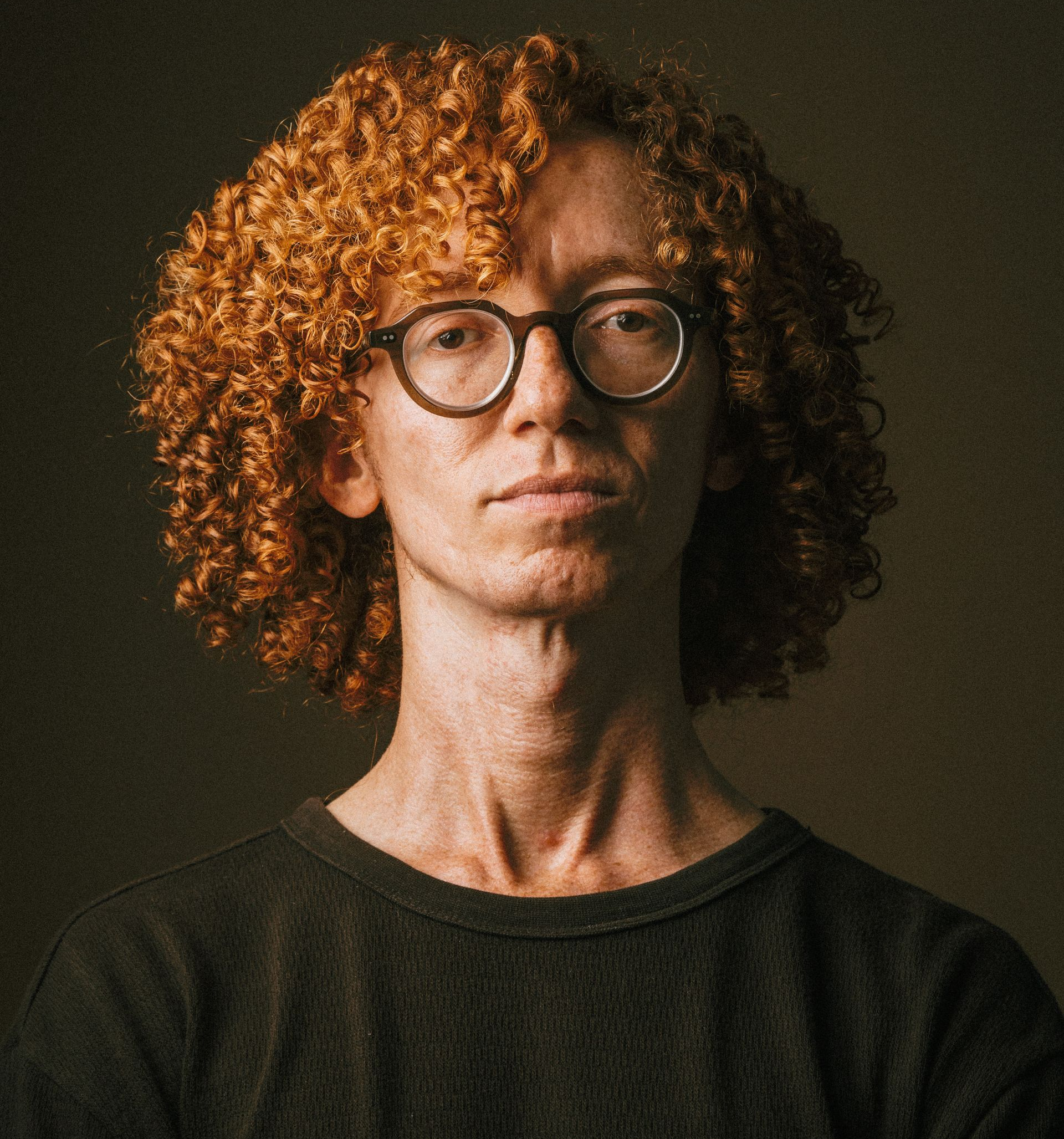 Retrato de una persona con cabello castaño rojizo rizado y gafas, que lleva un suéter oscuro sobre un fondo oscuro.