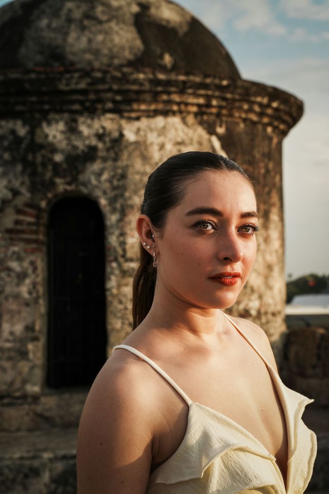 Mujer con vestido color crema de pie junto a una torre de piedra desgastada al atardecer.