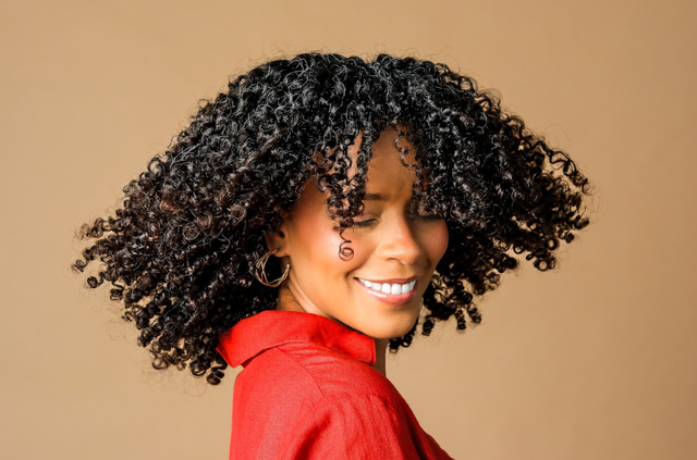 Mujer con cabello rizado y blusa roja sonriendo sobre un fondo beige.