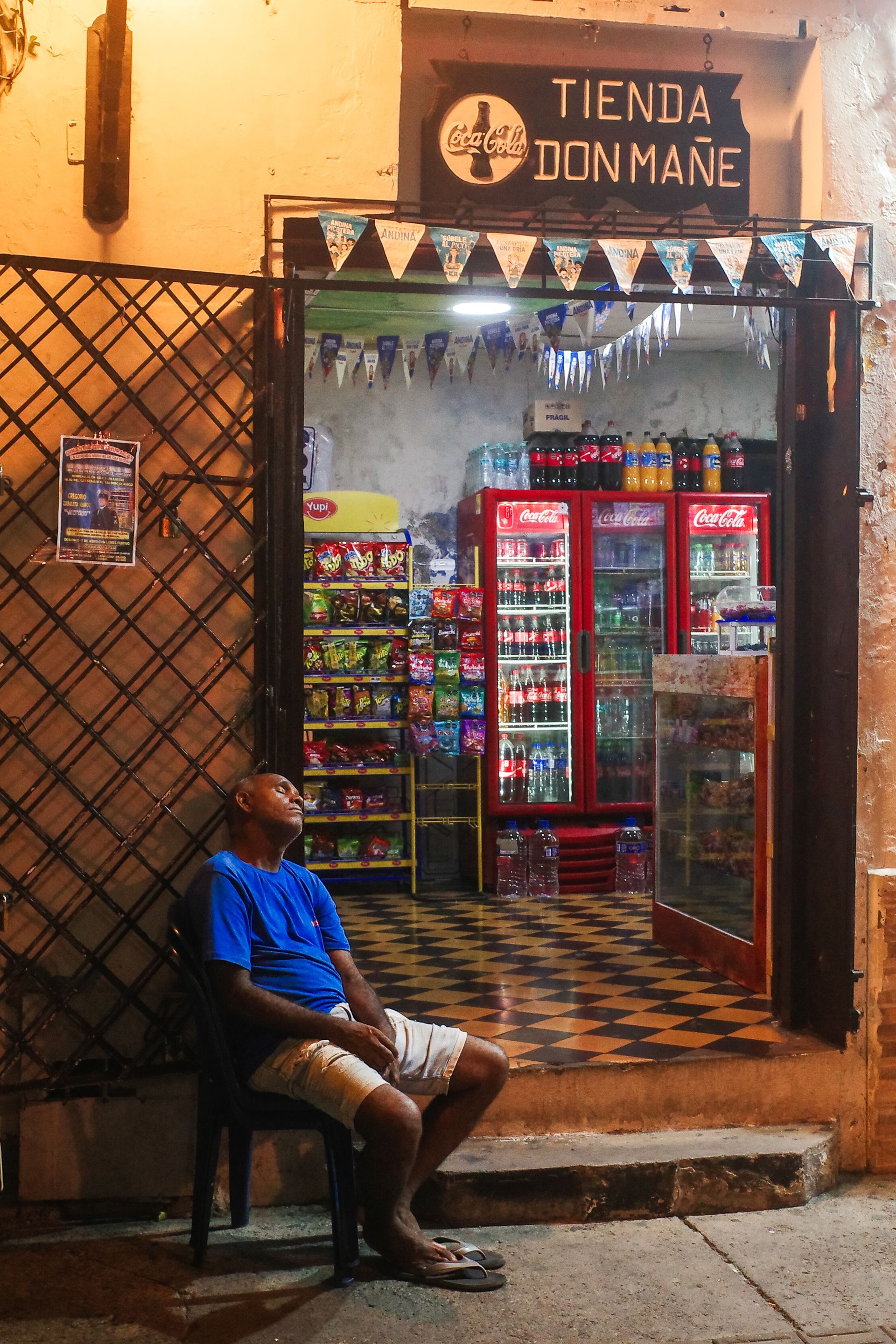 Entrada a una pequeña tienda con una nevera de puerta de cristal roja y una persona sentada