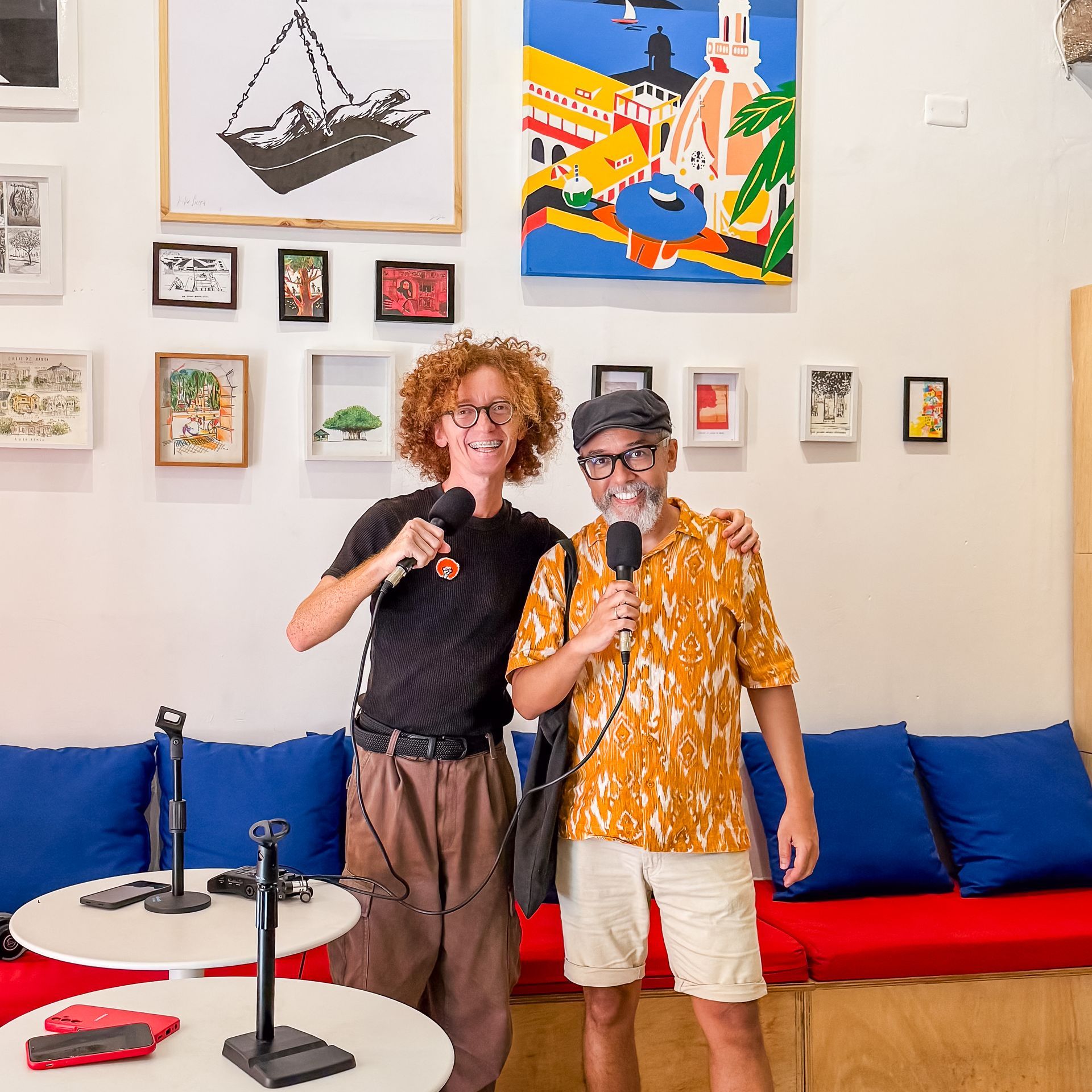 Dos personas cantando con micrófonos en una colorida sala de estar con cuadros en las paredes y un sofá azul.