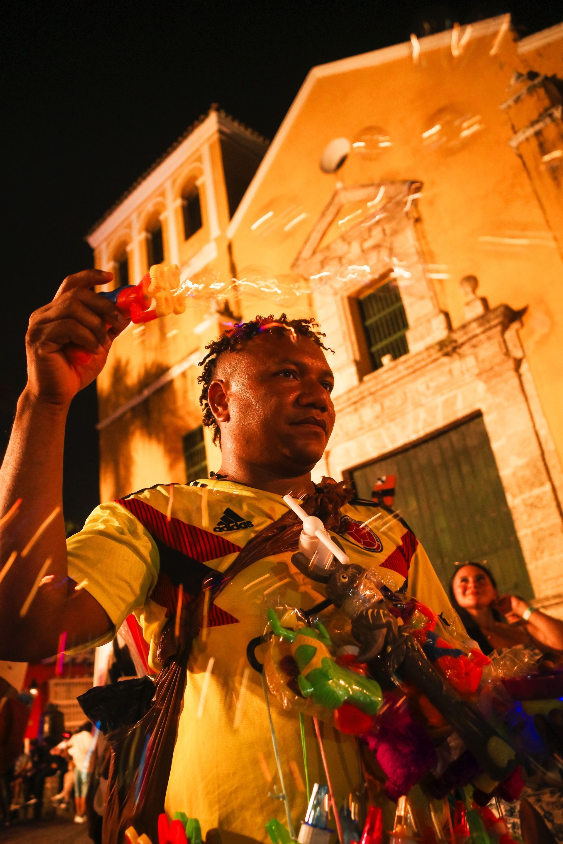 Persona con un disfraz colorido en una celebración callejera nocturna, sosteniendo un objeto rojo entre confeti.