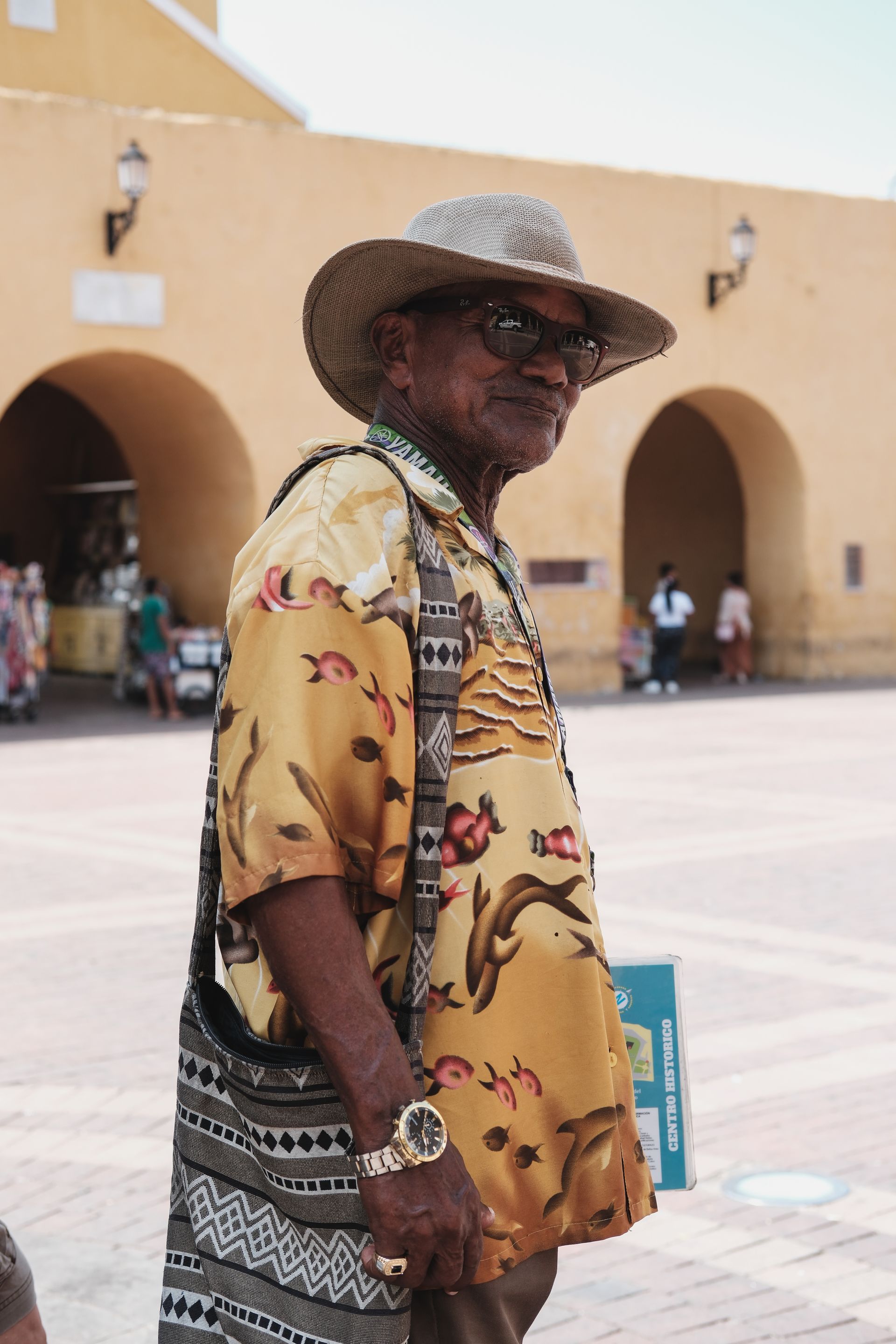 Hombre con camisa estampada y sombrero de paja de pie en un patio soleado con arcos.