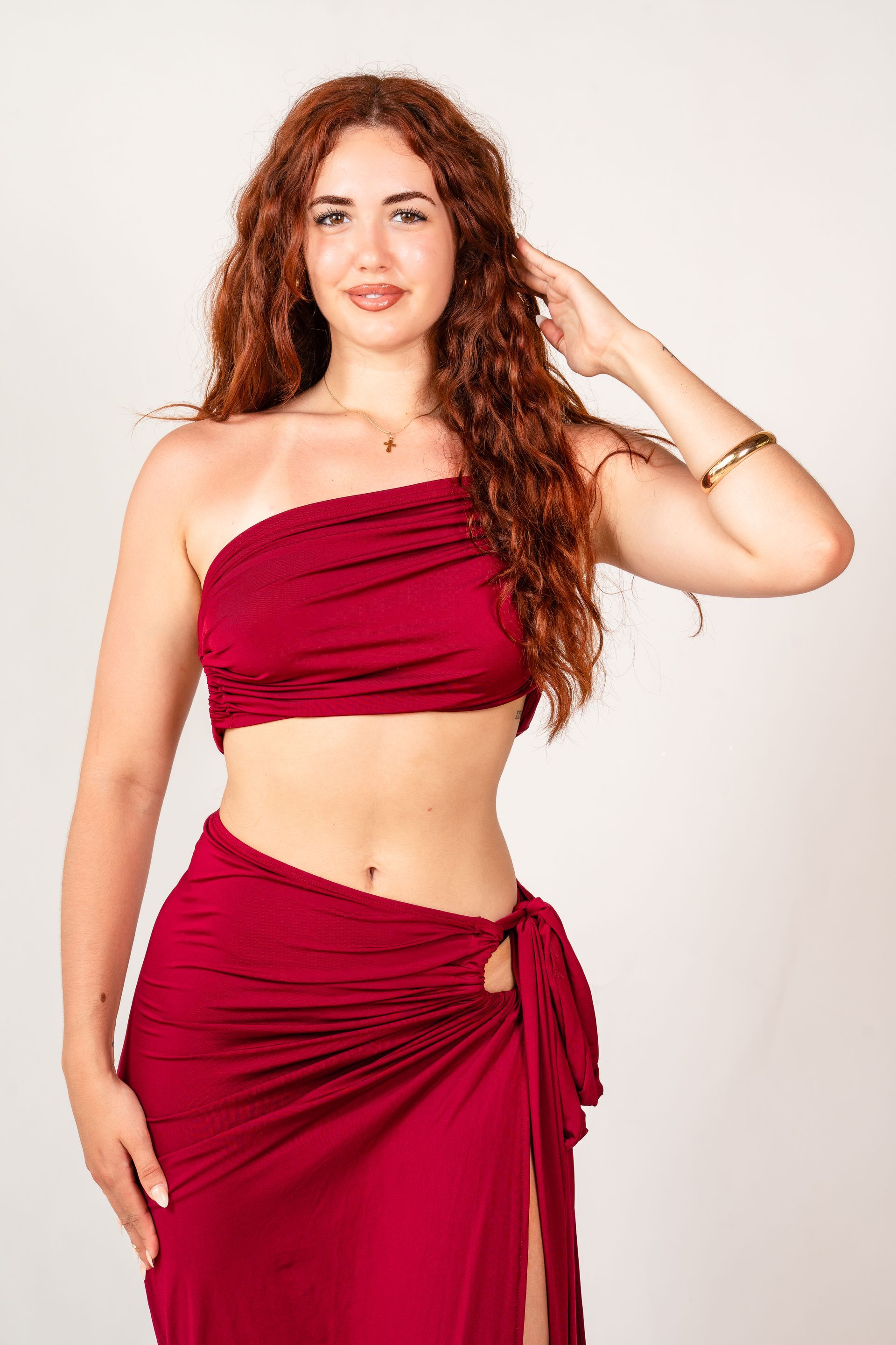 Mujer con top y falda rojos, de pie frente a un fondo claro con una mano en el cabello.