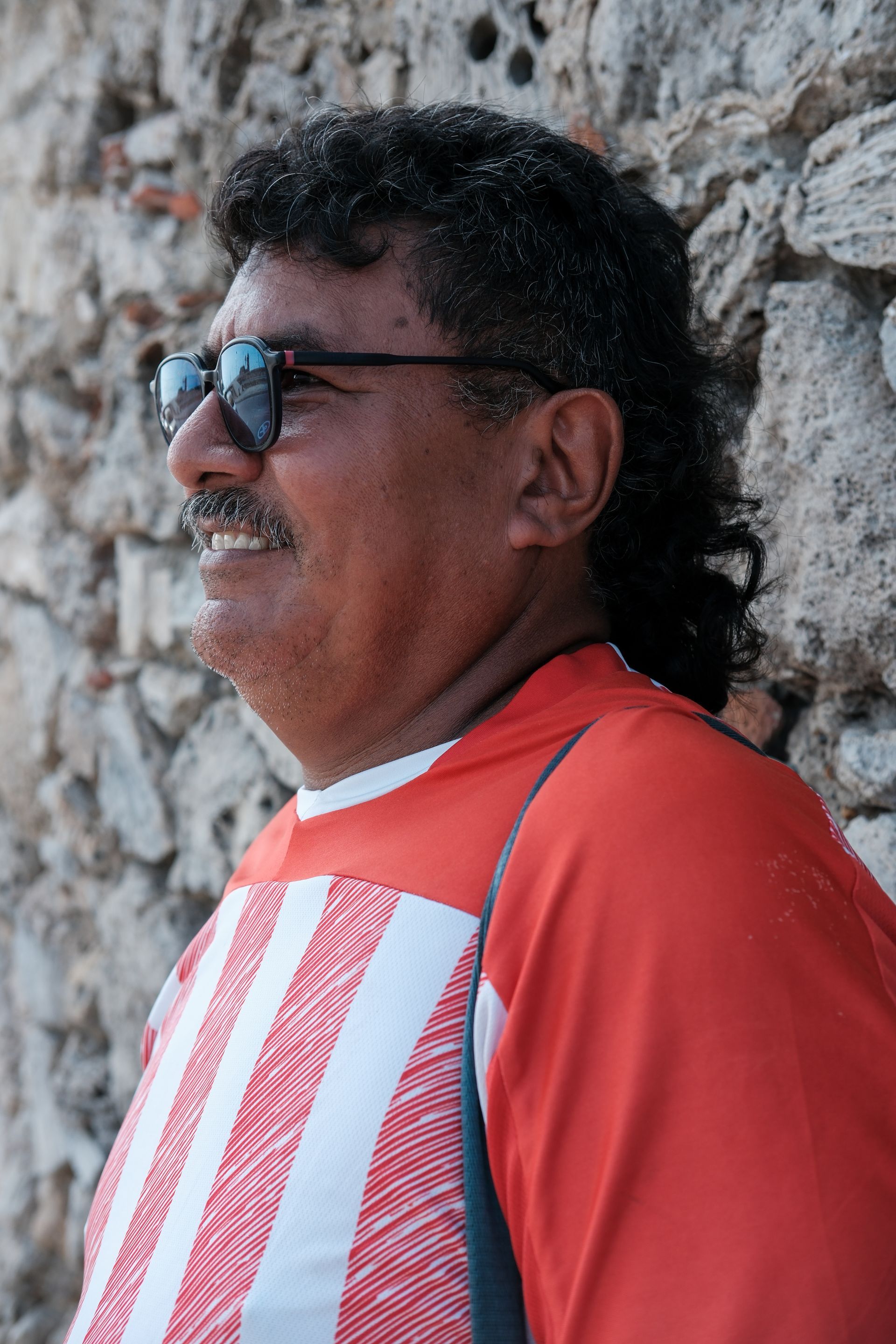 Hombre con camisa roja y blanca y gafas de sol, de pie junto a un muro de piedra.