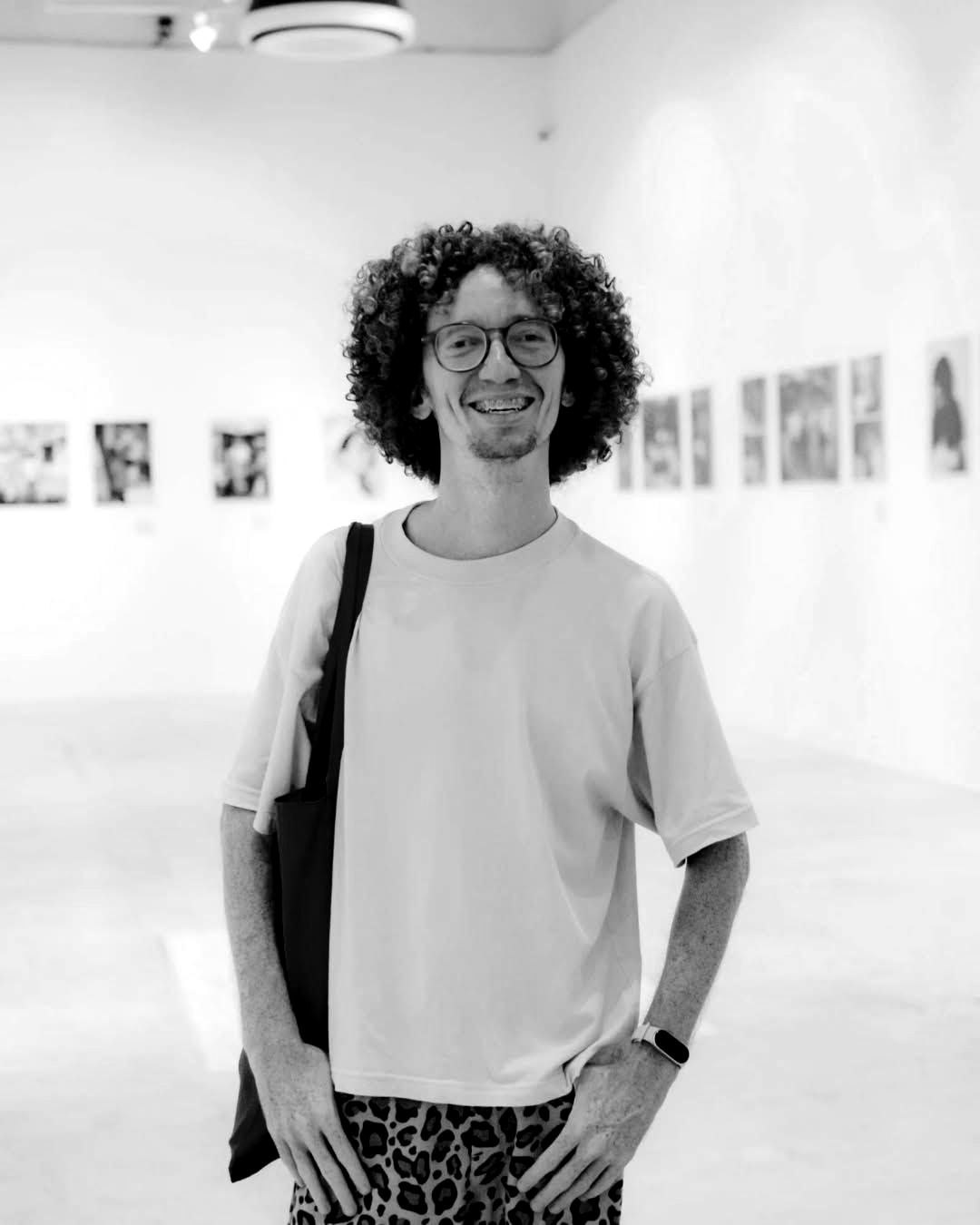 Persona con cabello rizado y gafas de pie en una luminosa galería de arte, sosteniendo un bolso al hombro y sonriendo.