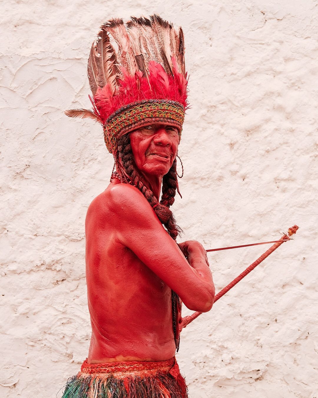 Persona con tocado ceremonial rojo y taparrabos, sosteniendo un arco, de pie contra una pared blanca.